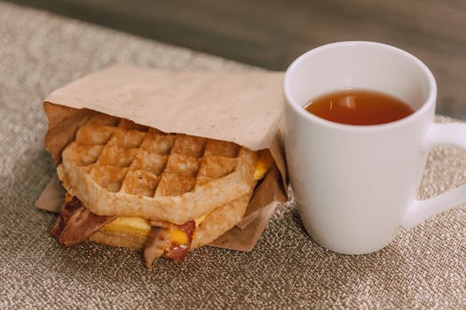 Tasty waffle sandwich with bacon and cheese next to a warm cup of tea.